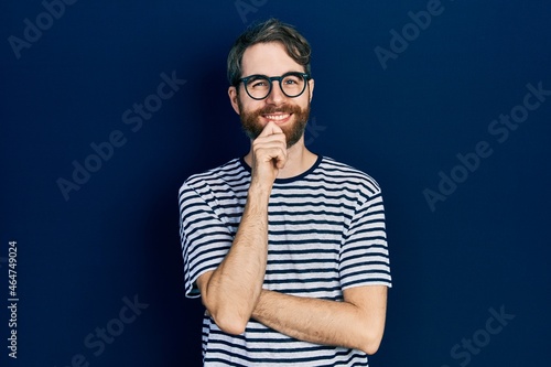 Caucasian man with beard wearing striped t shirt and glasses looking confident at the camera with smile with crossed arms and hand raised on chin. thinking positive.