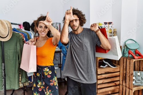 Young hispanic couple holding shopping bags at retail shop making fun of people with fingers on forehead doing loser gesture mocking and insulting.