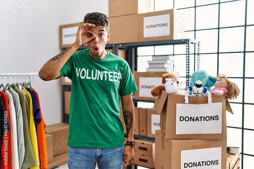 Young handsome hispanic man wearing volunteer t shirt at donations stand doing ok gesture shocked with surprised face, eye looking through fingers. unbelieving expression. © Krakenimages.com