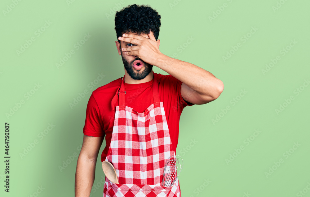 Young arab man with beard wearing cook apron peeking in shock covering face and eyes with hand, looking through fingers with embarrassed expression. Young arab man with beard wearing cook apron peeking in shock covering face and eyes with hand, looking through fingers with embarrassed expression.