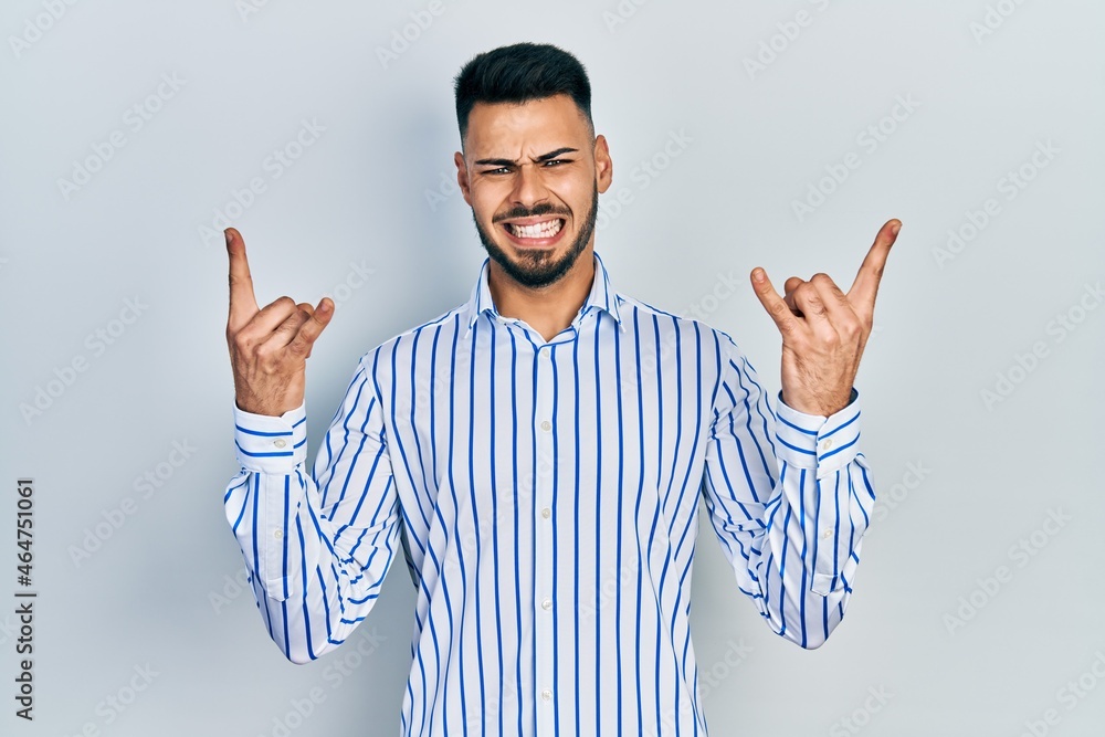 Naklejka premium Young hispanic man with beard wearing casual striped shirt shouting with crazy expression doing rock symbol with hands up. music star. heavy concept.