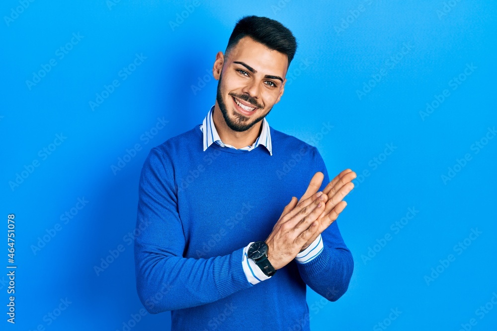 Young hispanic man with beard wearing casual blue sweater clapping and applauding happy and joyful, smiling proud hands together Young hispanic man with beard wearing casual blue sweater clapping and applauding happy and joyful, smiling proud hands together