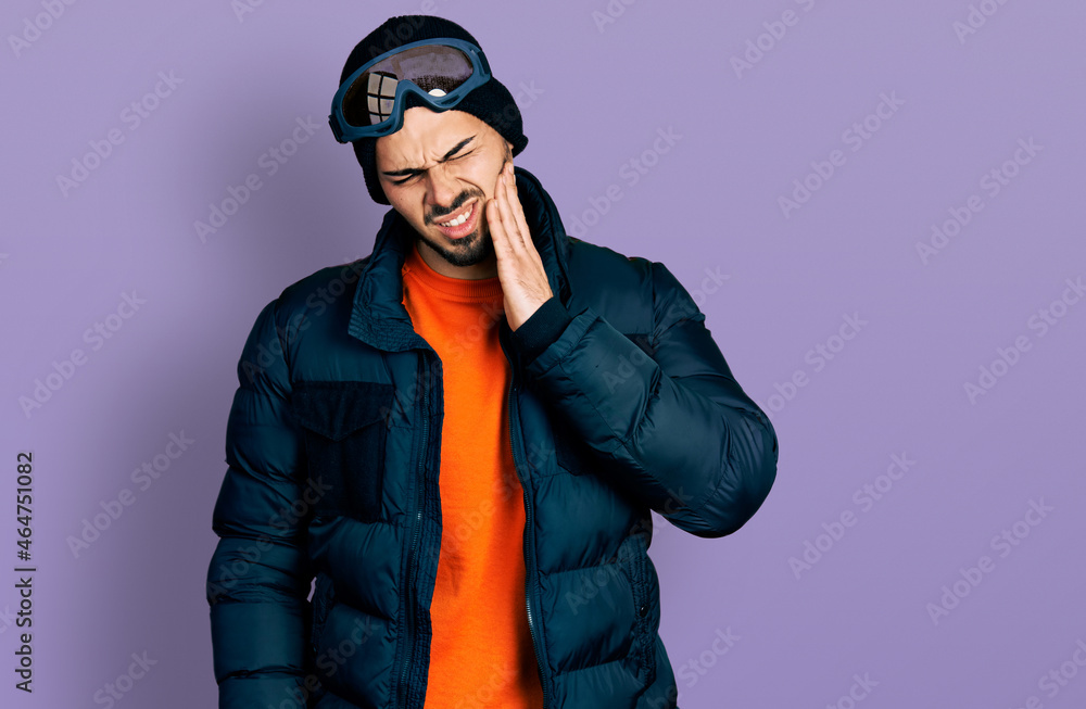 Young hispanic man with beard wearing snow wear and sky glasses touching mouth with hand with painful expression because of toothache or dental illness on teeth. dentist Young hispanic man with beard wearing snow wear and sky glasses touching mouth with hand with painful expression because of toothache or dental illness on teeth. dentist