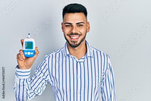 Young hispanic man with beard holding glucometer device looking positive and happy standing and smiling with a confident smile showing teeth photo