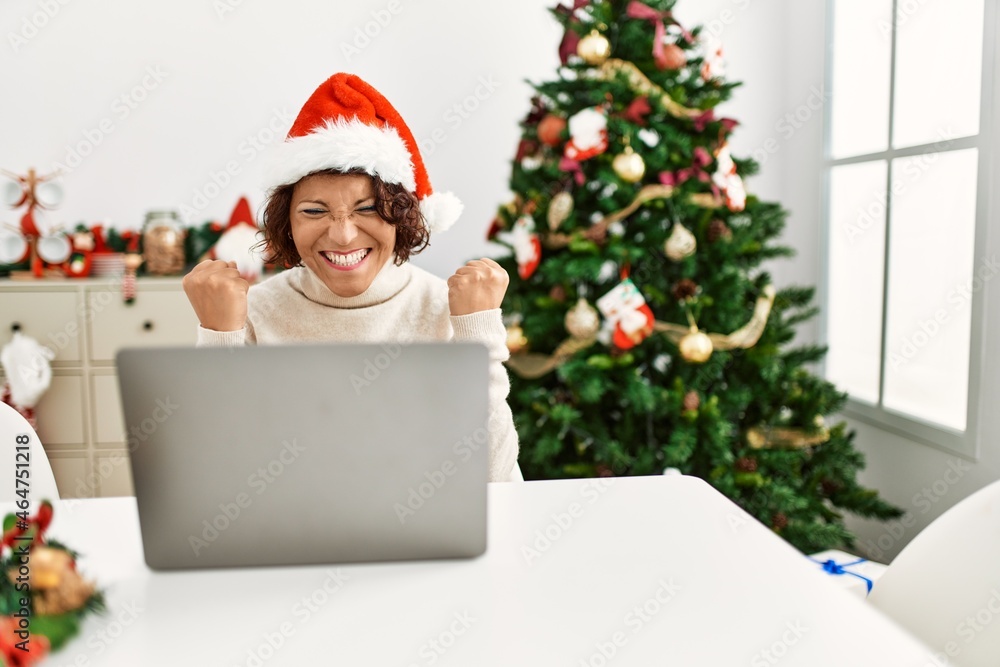 Middle age hispanic woman using laptop sitting by christmas tree screaming proud, celebrating victory and success very excited with raised arms