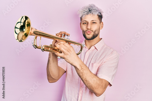 Young hispanic man with modern dyed hair playing trumpet looking at the camera blowing a kiss being lovely and sexy. love expression.