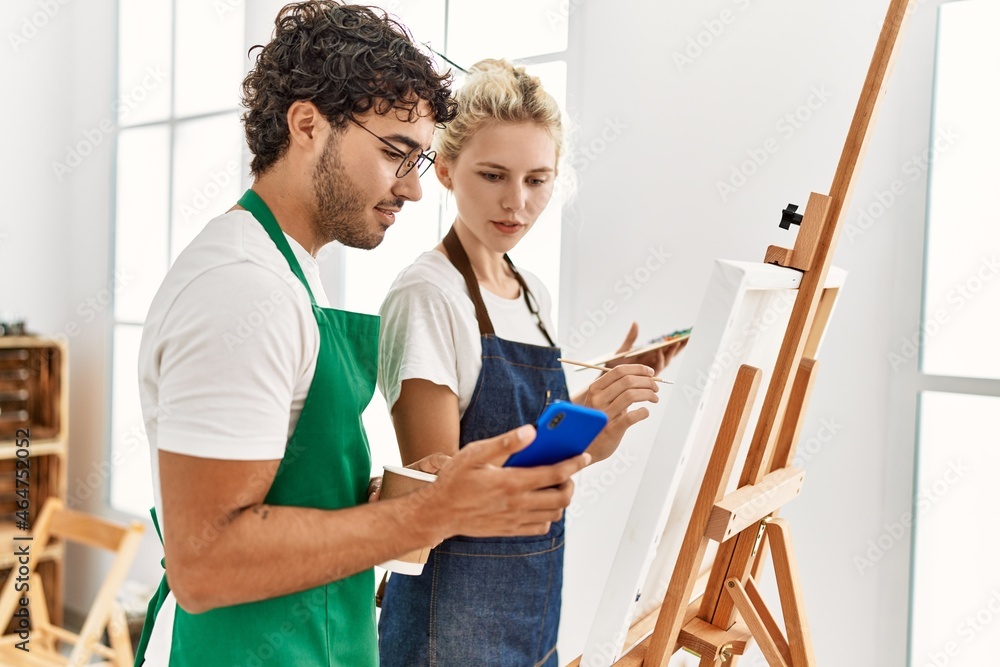 Young artist couple smiling happy using smartphone and painting at art studio. Young artist couple smiling happy using smartphone and painting at art studio.