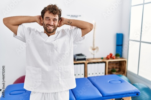 Young handsome physiotherapist man working at pain recovery clinic relaxing and stretching  arms and hands behind head and neck smiling happy
