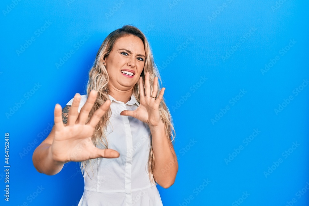 Beautiful young blonde woman wearing casual white shirt afraid and terrified with fear expression stop gesture with hands, shouting in shock. panic concept.