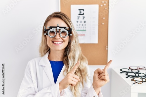 Young beautiful optician woman wearing optometry glasses at the clinic smiling and looking at the camera pointing with two hands and fingers to the side.