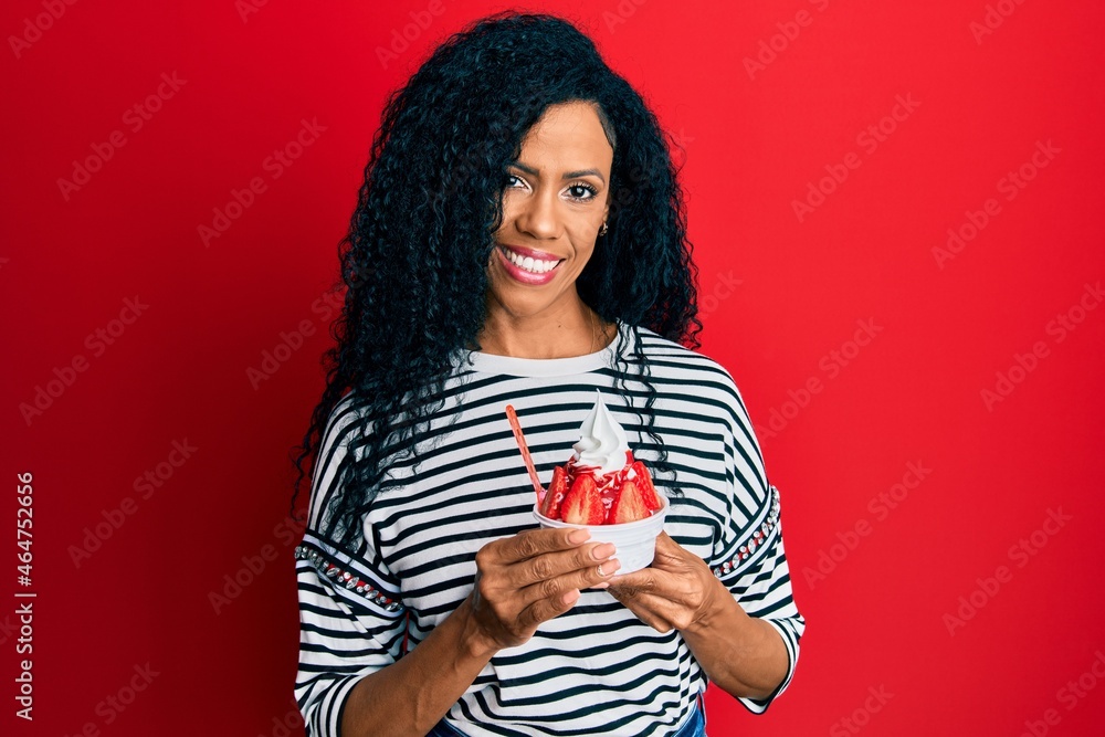 Middle age african american woman eating strawberry ice cream looking positive and happy standing and smiling with a confident smile showing teeth Middle age african american woman eating strawberry ice cream looking positive and happy standing and smiling with a confident smile showing teeth
