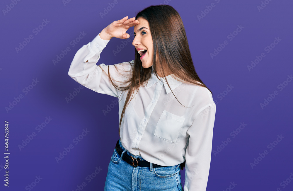 Young brunette teenager wearing business white shirt very happy and smiling looking far away with hand over head. searching concept. Young brunette teenager wearing business white shirt very happy and smiling looking far away with hand over head. searching concept.