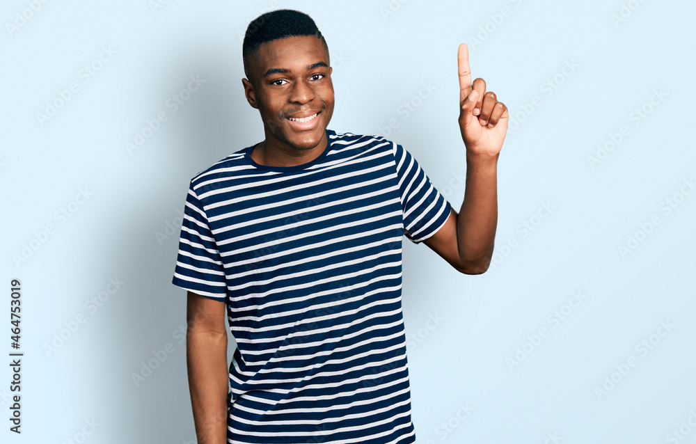 Fototapeta premium Young african american man wearing casual striped t shirt smiling with an idea or question pointing finger up with happy face, number one