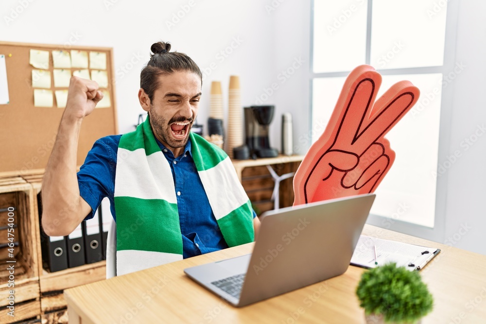Young hispanic man watching football on laptop cheering game at the office celebrating victory with happy smile and winner expression with raised hands Young hispanic man watching football on laptop cheering game at the office celebrating victory with happy smile and winner expression with raised hands
