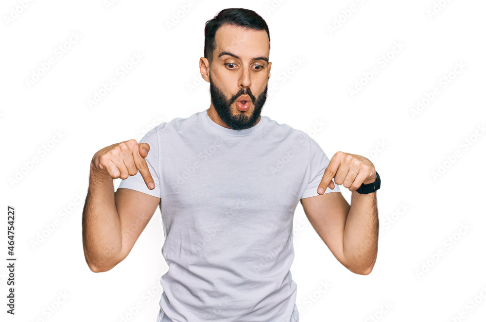 Young man with beard wearing casual white t shirt pointing down with fingers showing advertisement, surprised face and open mouth Young man with beard wearing casual white t shirt pointing down with fingers showing advertisement, surprised face and open mouth