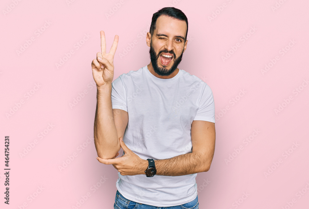 Young man with beard wearing casual white t shirt smiling with happy face winking at the camera doing victory sign with fingers. number two. Young man with beard wearing casual white t shirt smiling with happy face winking at the camera doing victory sign with fingers. number two.