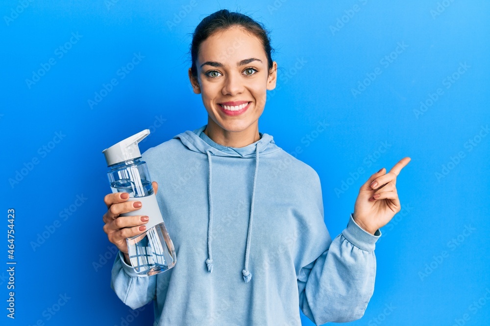 Young caucasian blonde woman drinking bottle of water smiling happy pointing with hand and finger to the side Young caucasian blonde woman drinking bottle of water smiling happy pointing with hand and finger to the side