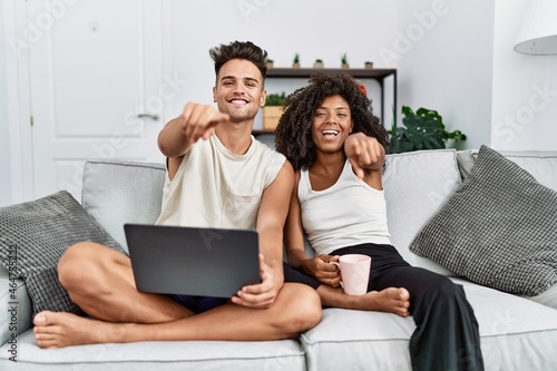 Young interracial couple using laptop at home sitting on the sofa pointing to you and the camera with fingers, smiling positive and cheerful