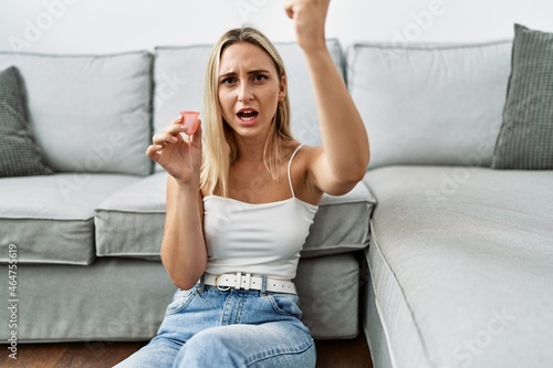 Young blonde woman holding menstrual cup annoyed and frustrated shouting with anger, yelling crazy with anger and hand raised
