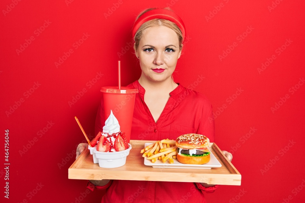 Young blonde woman eating a tasty classic burger with fries and soda relaxed with serious expression on face. simple and natural looking at the camera. Young blonde woman eating a tasty classic burger with fries and soda relaxed with serious expression on face. simple and natural looking at the camera.