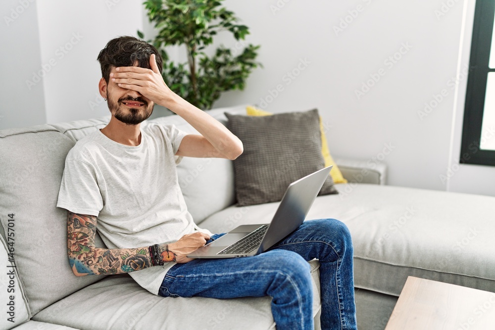 Hispanic man with beard sitting on the sofa smiling and laughing with hand on face covering eyes for surprise. blind concept.