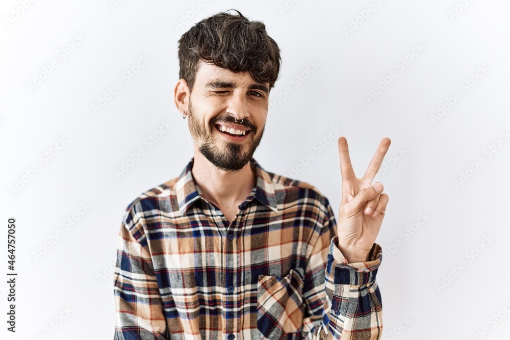 Hispanic man with beard standing over isolated background smiling with happy face winking at the camera doing victory sign. number two. Hispanic man with beard standing over isolated background smiling with happy face winking at the camera doing victory sign. number two.