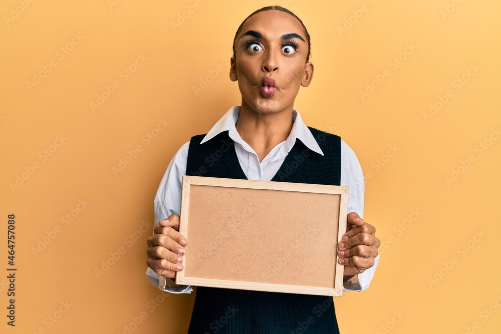 Hispanic man wearing make up and long hair holding empty corkboard making fish face with mouth and squinting eyes, crazy and comical. Hispanic man wearing make up and long hair holding empty corkboard making fish face with mouth and squinting eyes, crazy and comical.