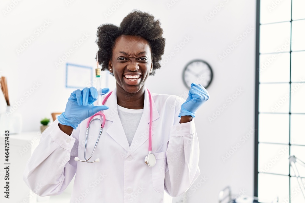 African doctor woman holding syringe at medical clinic screaming proud, celebrating victory and success very excited with raised arm African doctor woman holding syringe at medical clinic screaming proud, celebrating victory and success very excited with raised arm