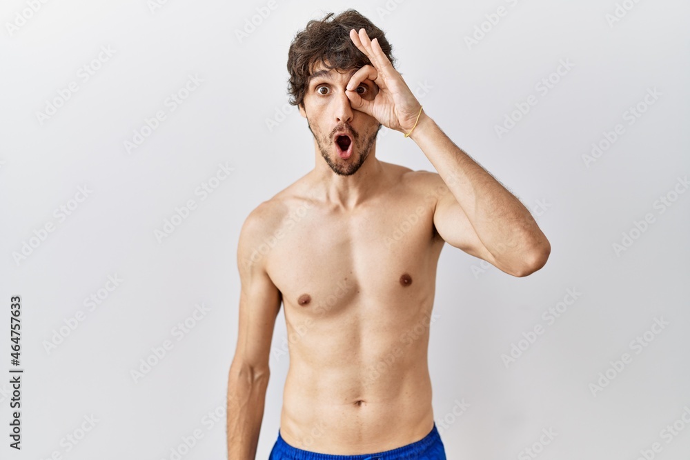 Fototapeta premium Young hispanic man standing shirtless over isolated, background doing ok gesture shocked with surprised face, eye looking through fingers. unbelieving expression.