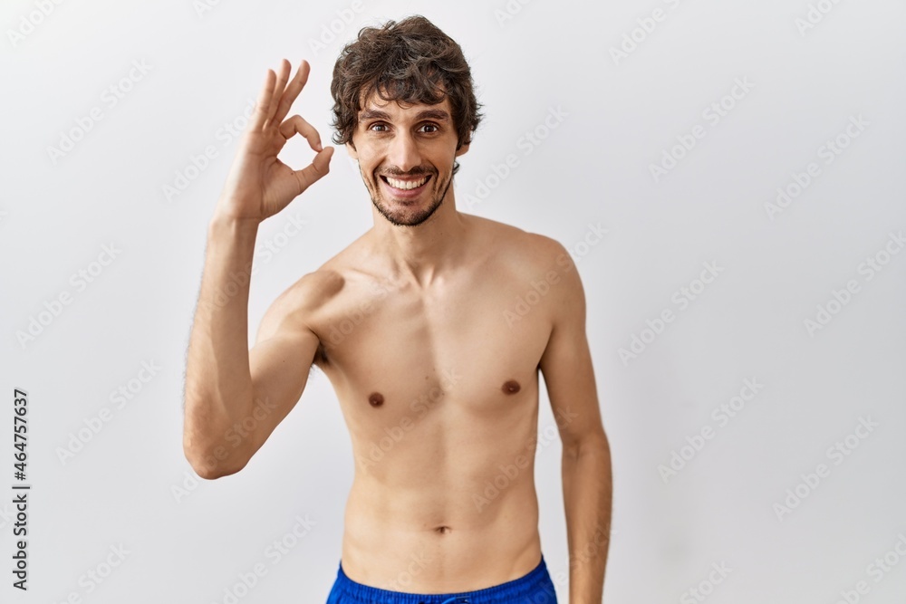 Fototapeta premium Young hispanic man standing shirtless over isolated, background smiling positive doing ok sign with hand and fingers. successful expression.