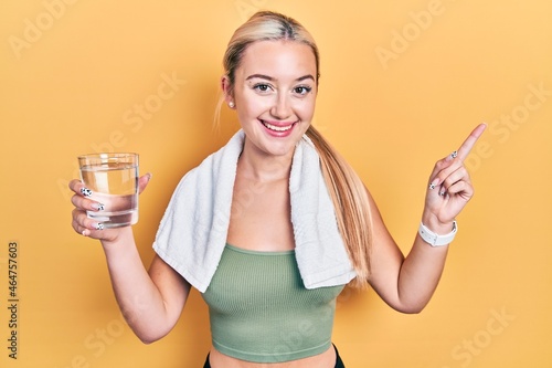 Young blonde girl wearing sportswear drinking glass of water smiling happy pointing with hand and finger to the side © Krakenimages.com