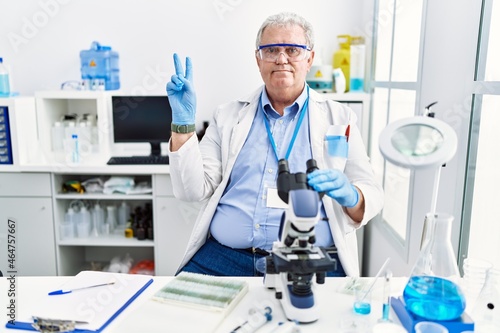 Senior caucasian man working at scientist laboratory smiling with happy face winking at the camera doing victory sign with fingers. number two.