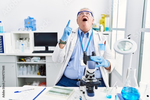 Senior caucasian man working at scientist laboratory amazed and surprised looking up and pointing with fingers and raised arms.