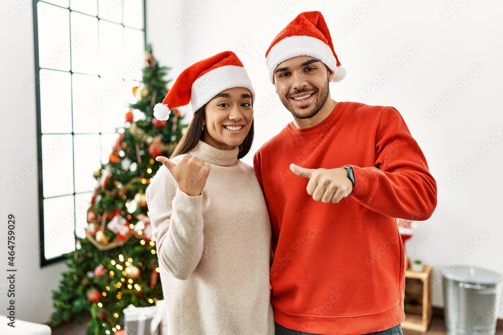 Young hispanic couple standing by christmas tree pointing to the back behind with hand and thumbs up, smiling confident Young hispanic couple standing by christmas tree pointing to the back behind with hand and thumbs up, smiling confident