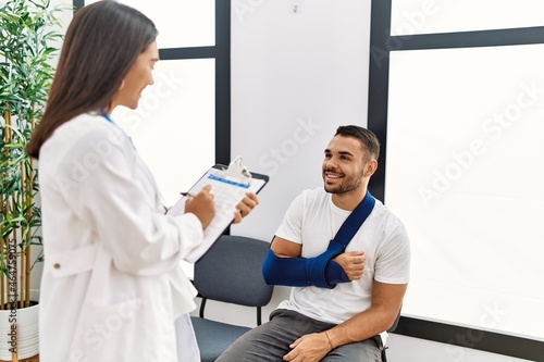 Youg latin injuried man wearing sling talking with the doctor at clinic waiting room.