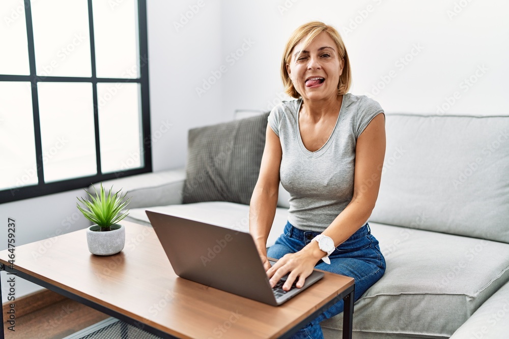 Middle age blonde woman using laptop at home sticking tongue out happy with funny expression. emotion concept. Middle age blonde woman using laptop at home sticking tongue out happy with funny expression. emotion concept.