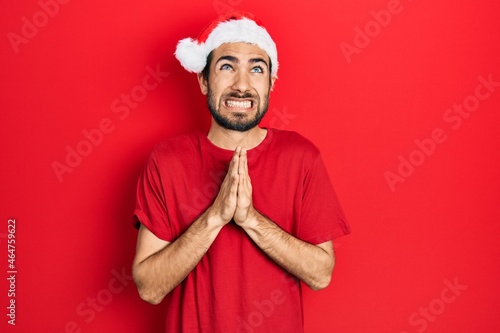 Young hispanic man wearing christmas hat begging and praying with hands together with hope expression on face very emotional and worried. begging.