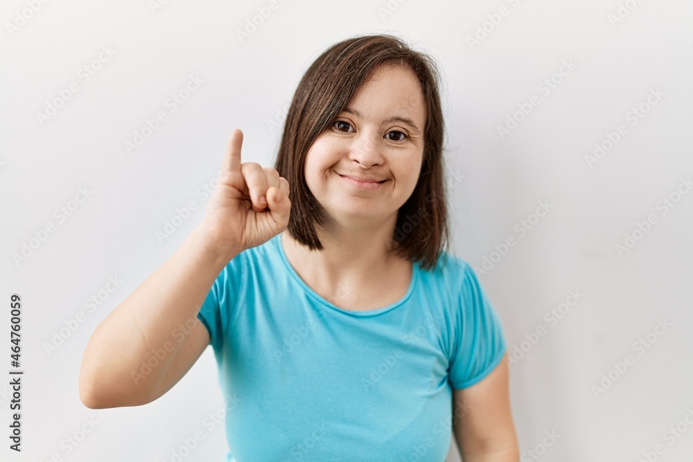 Young down syndrome woman standing over isolated background showing and pointing up with finger number one while smiling confident and happy. Young down syndrome woman standing over isolated background showing and pointing up with finger number one while smiling confident and happy.