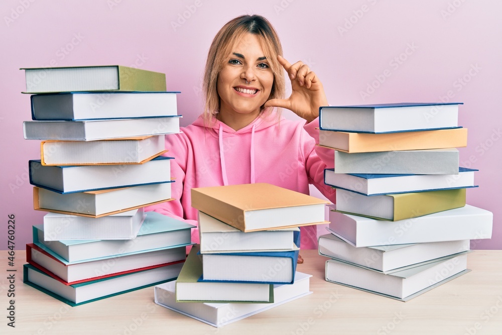 Young caucasian woman sitting on the table with books smiling and confident gesturing with hand doing small size sign with fingers looking and the camera. measure concept. Young caucasian woman sitting on the table with books smiling and confident gesturing with hand doing small size sign with fingers looking and the camera. measure concept.