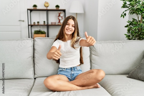 Young brunette teenager sitting on the sofa at home approving doing positive gesture with hand, thumbs up smiling and happy for success. winner gesture.