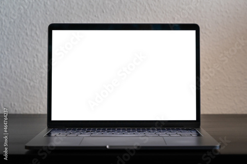 Empty laptop screen perfect for testing and a mockup of a design. Bright white screen in a dark environment. Device standing on a black wooden desk in front of a white wall.