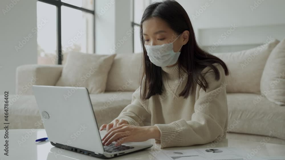 A thoughtful Asian woman in a medical mask using a laptop is working in the living room. Student studying online, distance learning A thoughtful Asian woman in a medical mask using a laptop is working in the living room. Student studying online, distance learning