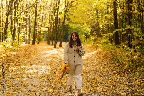 A young woman in a long shirt with a thermo mug in her hands walks through the autumn forest. Natural landscape. Beautiful forest. Autumn day.