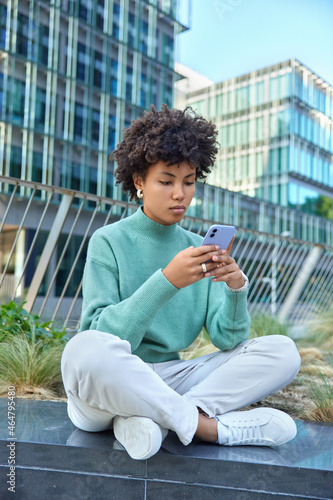 Concentrated serious young woman sits in lotus pose sends text messages and reads information on mobile phone watches video online uses smartphone technology poses utdoors against city buildings photo