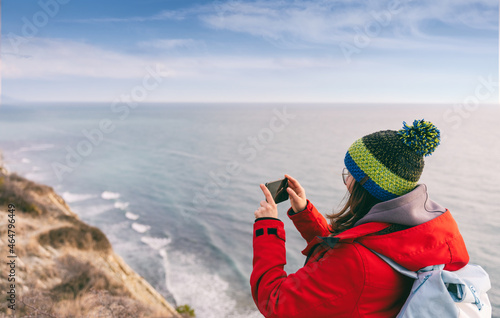 Young woman traveler in a jacket and a hat takes pictures of sea on a smartphone in cold weather