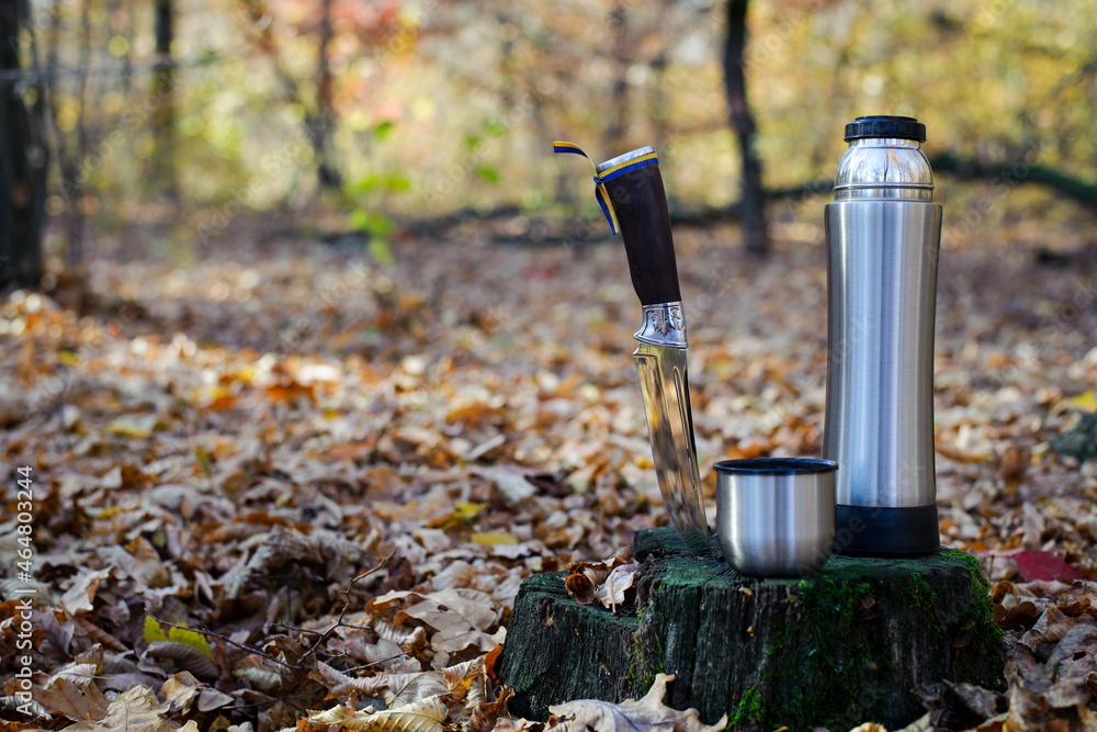Thermos and hunting knife. The concept of tourism, survival and hunting in the autumn in forest. a thermos of tea, a cup and a knife, on an old tree stump. dry leaves in the park. rest at nature.