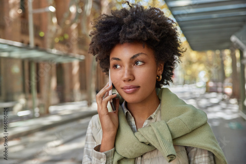 Generation communication concept. Headshot of thoughtful woman with curly hair talks with friend via mobile phone looks away wears casual clothes strolls outdoors uses international roaming connection