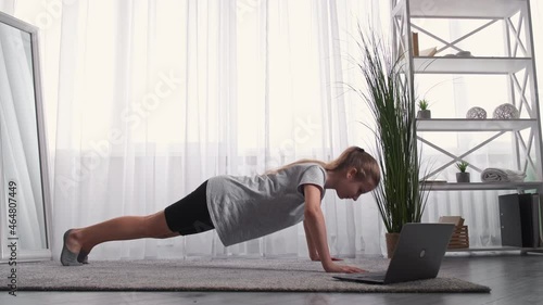 Online fitness. Teenager gym. Home training. Young girl doing press-ups workout looking laptop in light room interior.