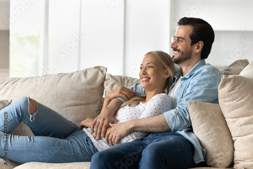 Happy bonding dreamy young family couple resting on cozy sofa, visualizing future looking in distance, planning vacation or enjoying leisure weekend moment together at home, good relations concep.t