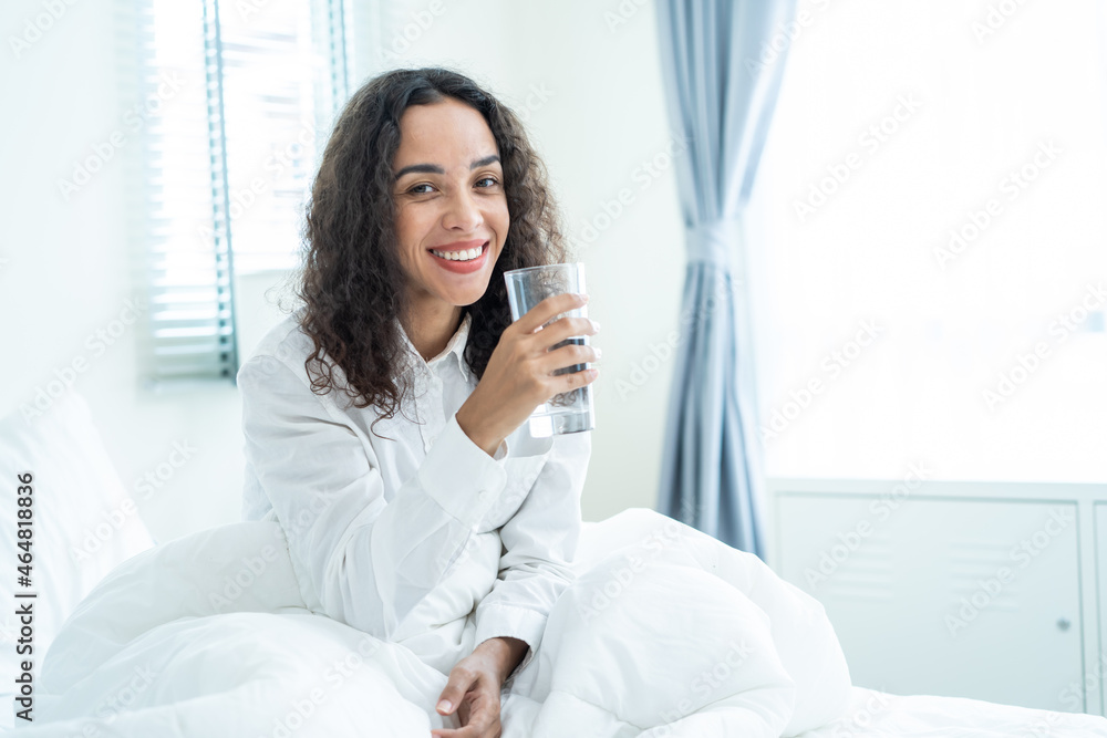 Portrait of Latino woman sit on bed, hold a glass of water in bedroom.